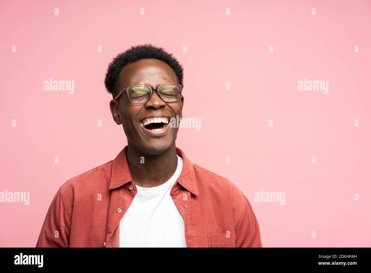 Laughing positive young Afro American man in red shirt in good mood, poses at camera with closed eyes. Overjoyed Black male in spectacles shows her br Stock Photo