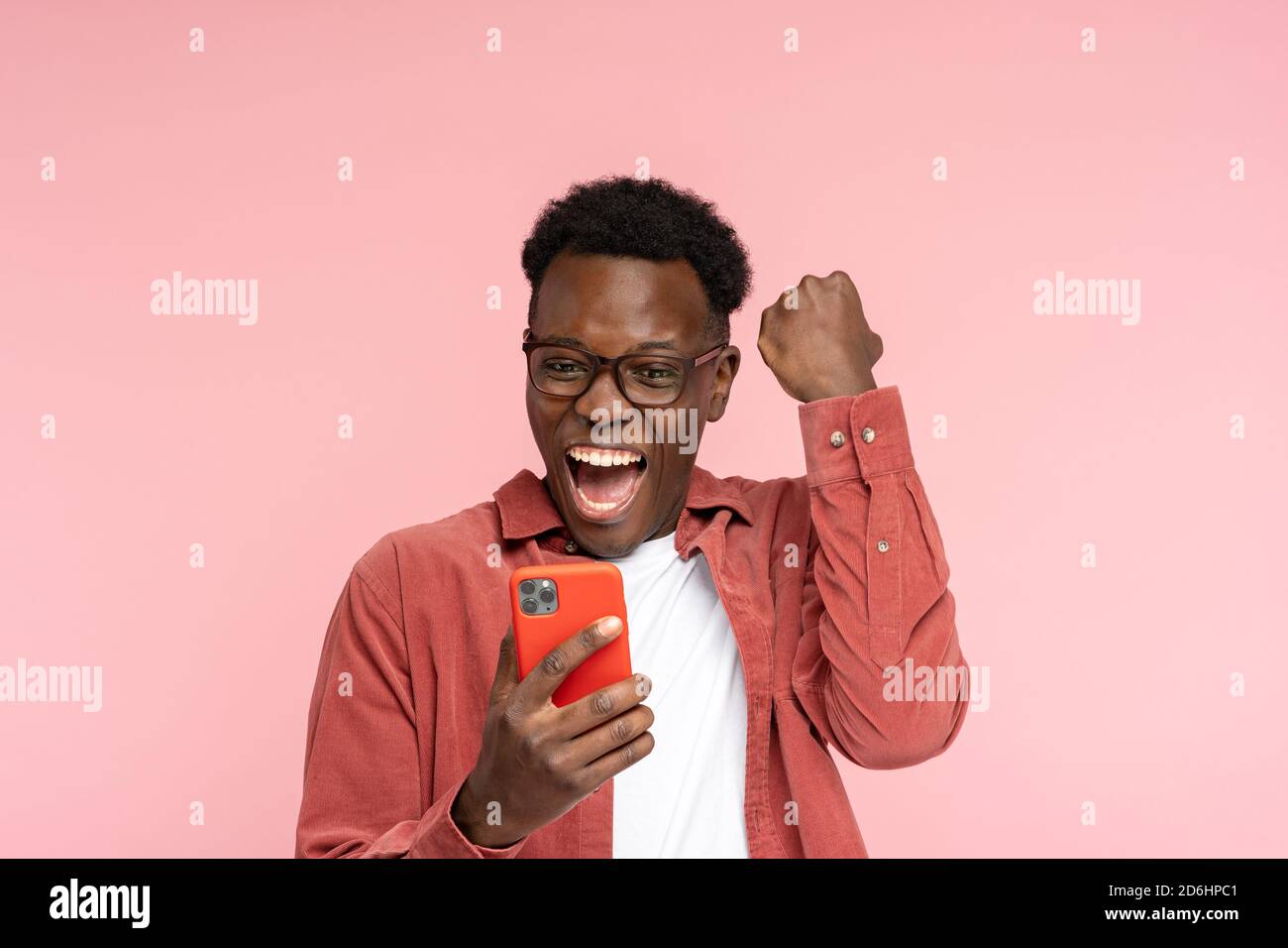 Excited Afro-American millennial man in glasses hold mobile phone isolated on pink studio background. Overjoyed black man look at smartphone, smile fe Stock Photo