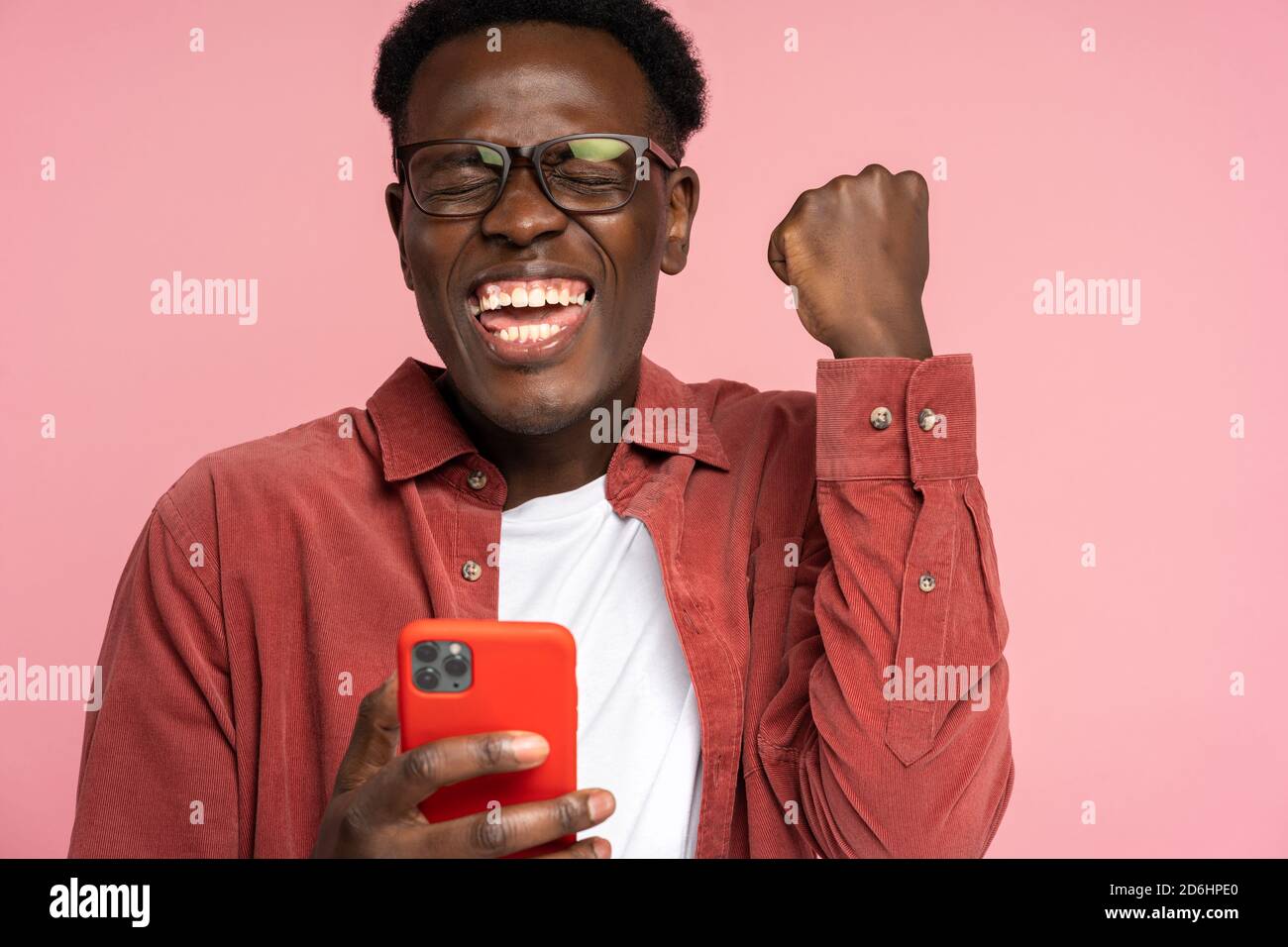 Excited Afro-American millennial man in glasses hold mobile phone isolated on pink studio background. Overjoyed black man look at smartphone, smile fe Stock Photo