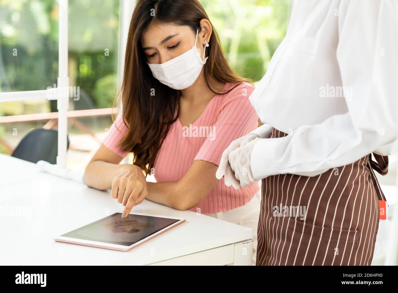 Asian waitress wear facemask and face shield using tablet to show restaurant electronic menu and recommend to customer. New normal technology and life Stock Photo