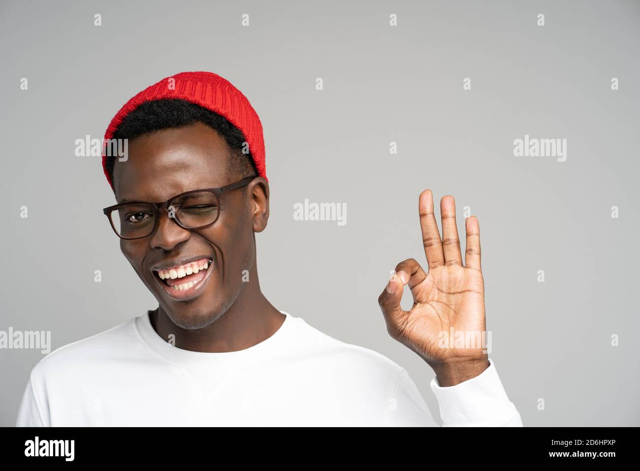 Cheerful playful young Afro American man wear red hat in good mood smiling broadly, winking, showing okay gesture over studio grey background. Positiv Stock Photo