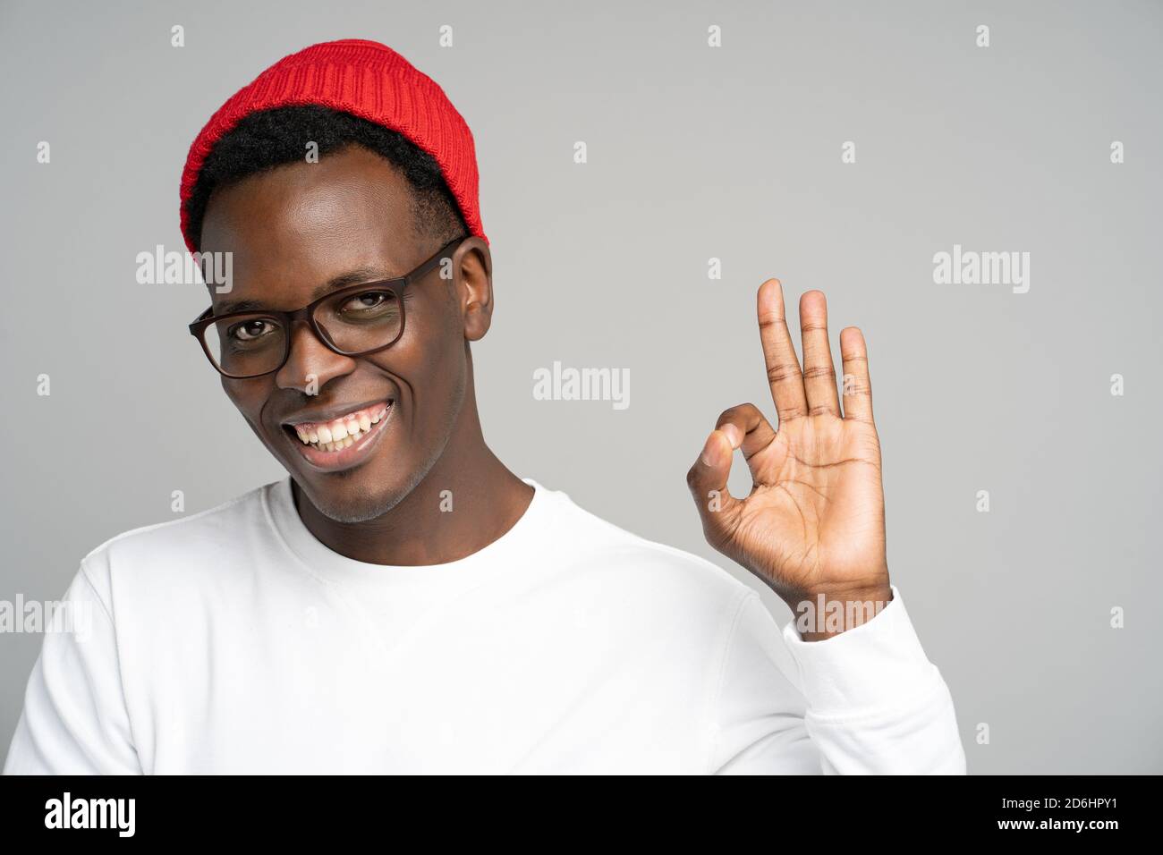 Cheerful playful young Afro American man wear red hat in good mood smiling broadly, showing okay gesture over studio grey background. Positive Black m Stock Photo