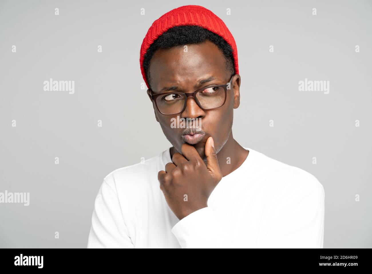 Studio portrait of unsure doubtful Black man in red hat wear glasses holds chin, looking right doubtfully, plans or reconsiders something, feeling hes Stock Photo