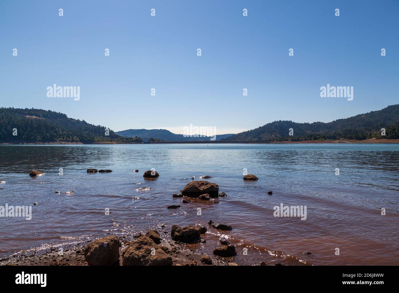 Looking across the surface of Lost Creek Lake to the William L. Jess Dam with a rocky foreground and the Cascade Mountains in the distance. Stock Photo