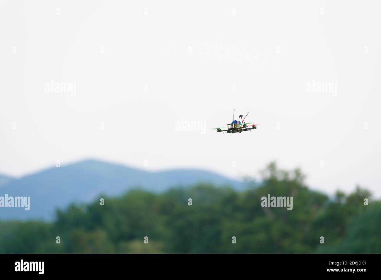 Colorful propellers lift this racing FPV quad copter drone through the air. Plain white sky background with blurred hills and trees beneath Stock Photo