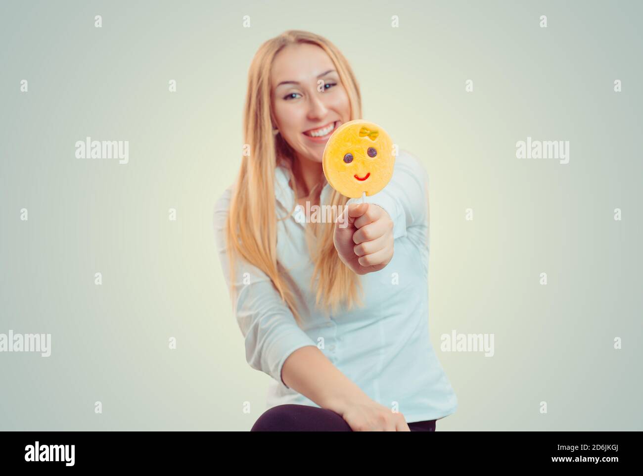 Bright blond woman holding giving stick with emoji lollipop smiling at camera isolated on light green yellow background with copy space. Blur on the f Stock Photo