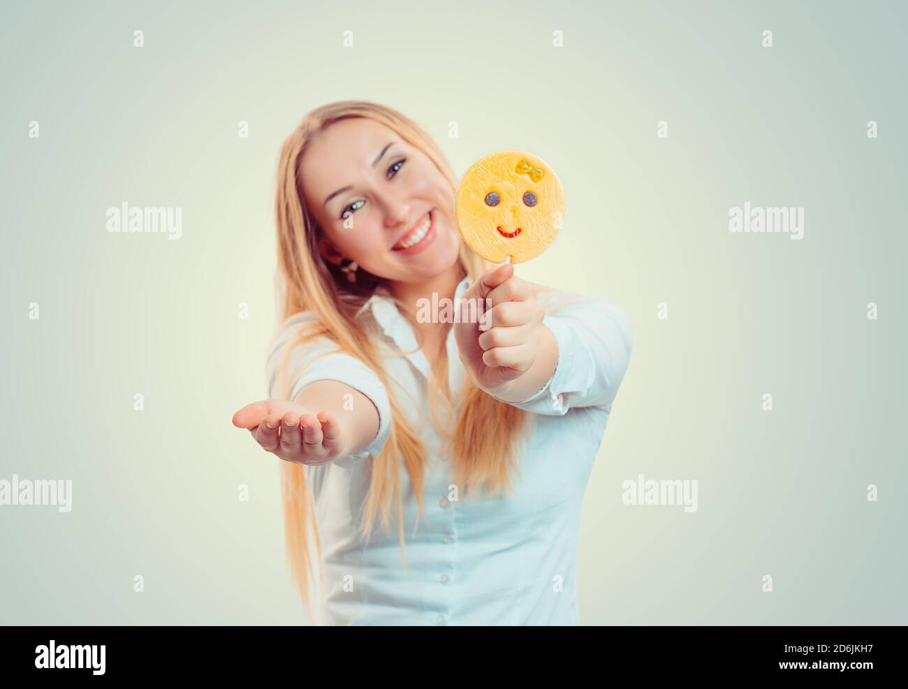Bright blond woman proposing giving candy stick with emoji lollipop smiling at camera isolated on light green yellow background with copy space. Blur Stock Photo