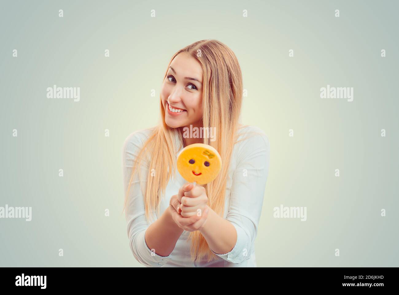 Bright blond woman holding giving proposing looking at you camera a stick, candy emoticon with emoji lollipop smiling at camera isolated on light gree Stock Photo