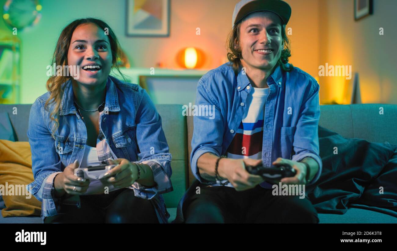 Excited Black Gamer Girl and Young Man Sitting on a Couch and Playing Video Games on Console. They Plays with Wireless Controllers. Cozy Room is Lit Stock Photo