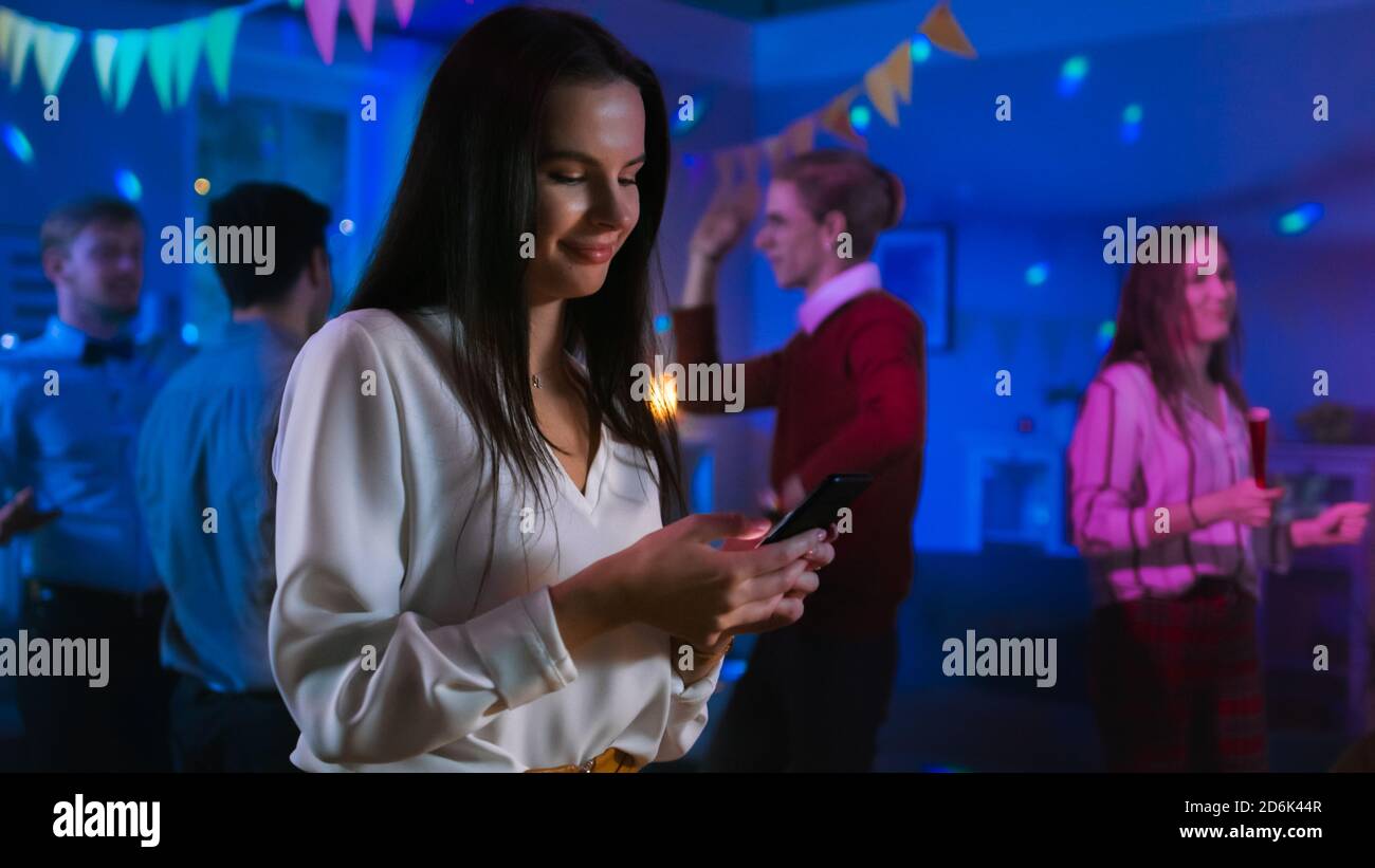 At the Wild House Party: Beautiful Young Girl Uses Smartphone, Browses  Through Internet and Social Network. In the Background Crowd of Young  People Stock Photo - Alamy