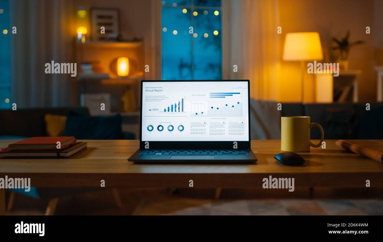 Laptop Computer Showing Statistical Infographics Stands on a Desk in the Living Room. In the Background Cozy Living Room with Warm Lights on. Stock Photo