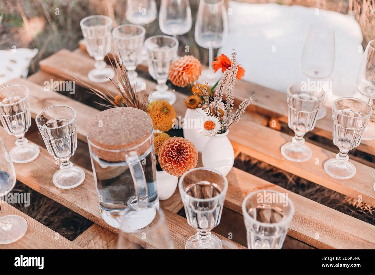 Set of elegant glassware placed on plank wooden table with floral arrangement on grassy meadow during festive autumn picnic Stock Photo
