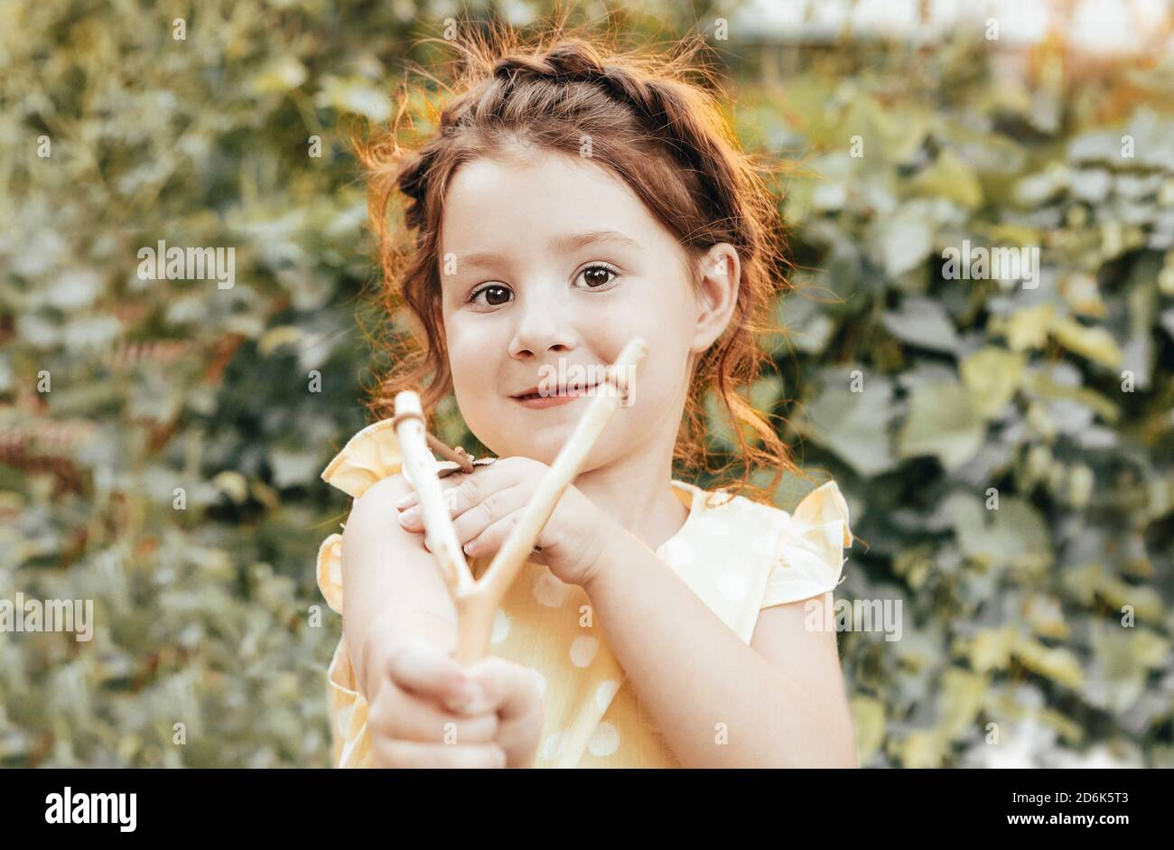 Cute naughty redhead little girl with braided hair aiming with slingshot at camera while spending summer day in garden Stock Photo