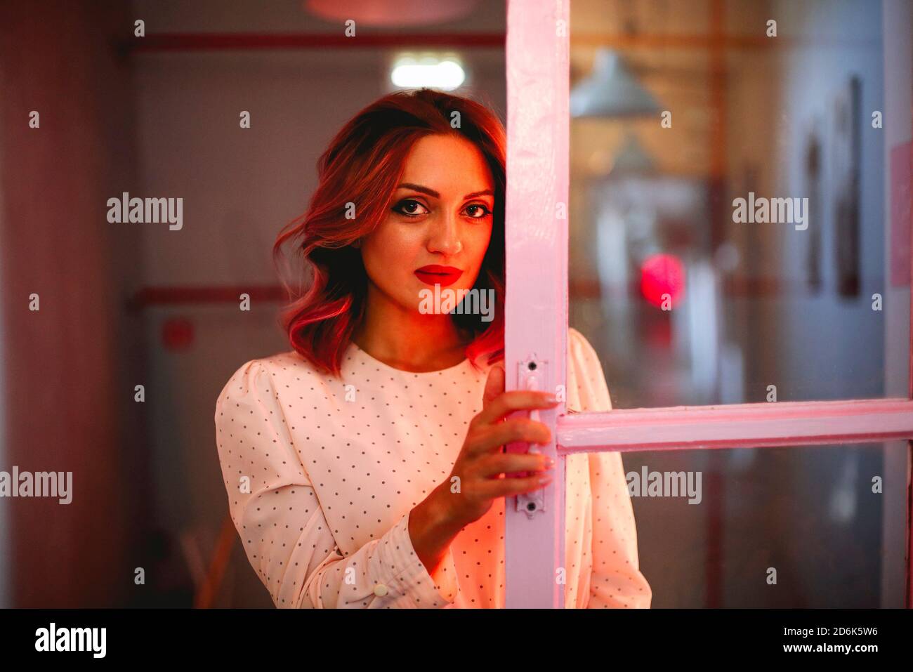 Charming female with wavy hair standing near door in room illuminated by red light and looking at camera Stock Photo