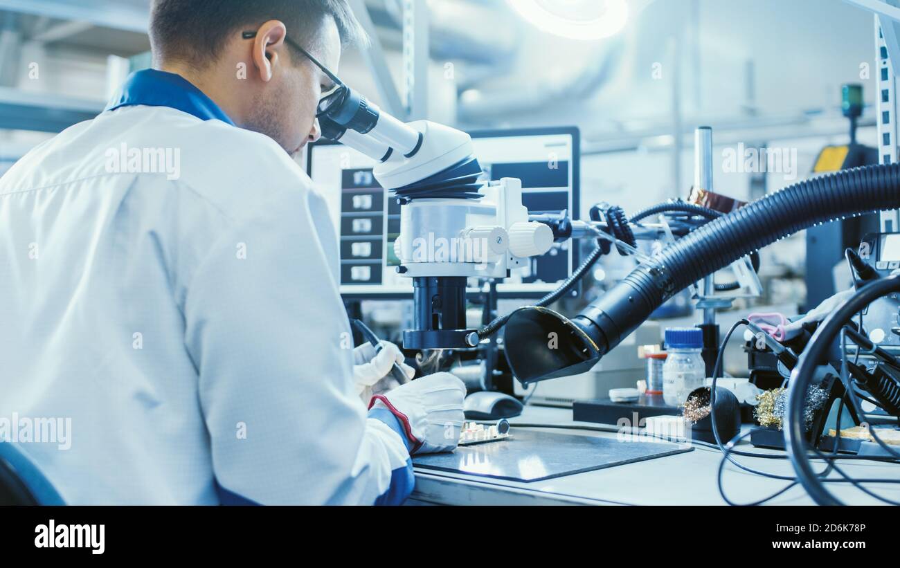 Electronics Factory Worker in White Work Coat is Soldering a Printed Circuit Board Through a Digital Microscope. High Tech Factory Facility. Stock Photo