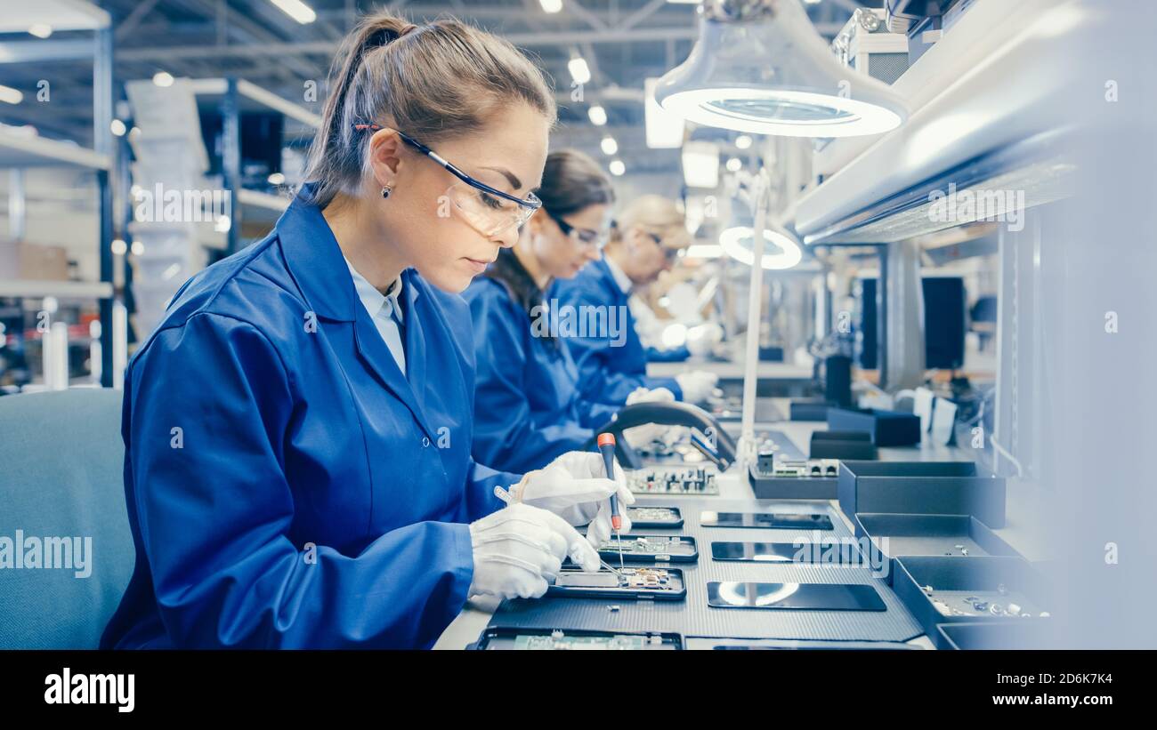 Woman Electronics Factory Worker in Blue Work Coat and Protective Glasses is Assembling Smartphones with Screwdriver. High Tech Factory Facility with Stock Photo