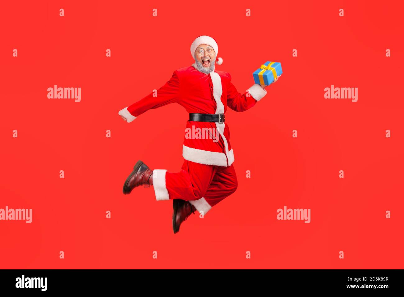 Extremely happy santa claus holding gift box jumping high and looking at camera with toothy smile, satisfied with winter holidays. Indoor studio shot Stock Photo