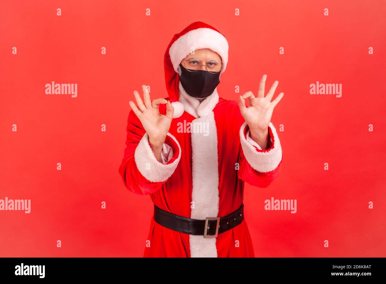 Positive optimistic man in santa claus costume with protective face mask showing ok gesture with fingers, cheering up on quarantine. Indoor studio sho Stock Photo