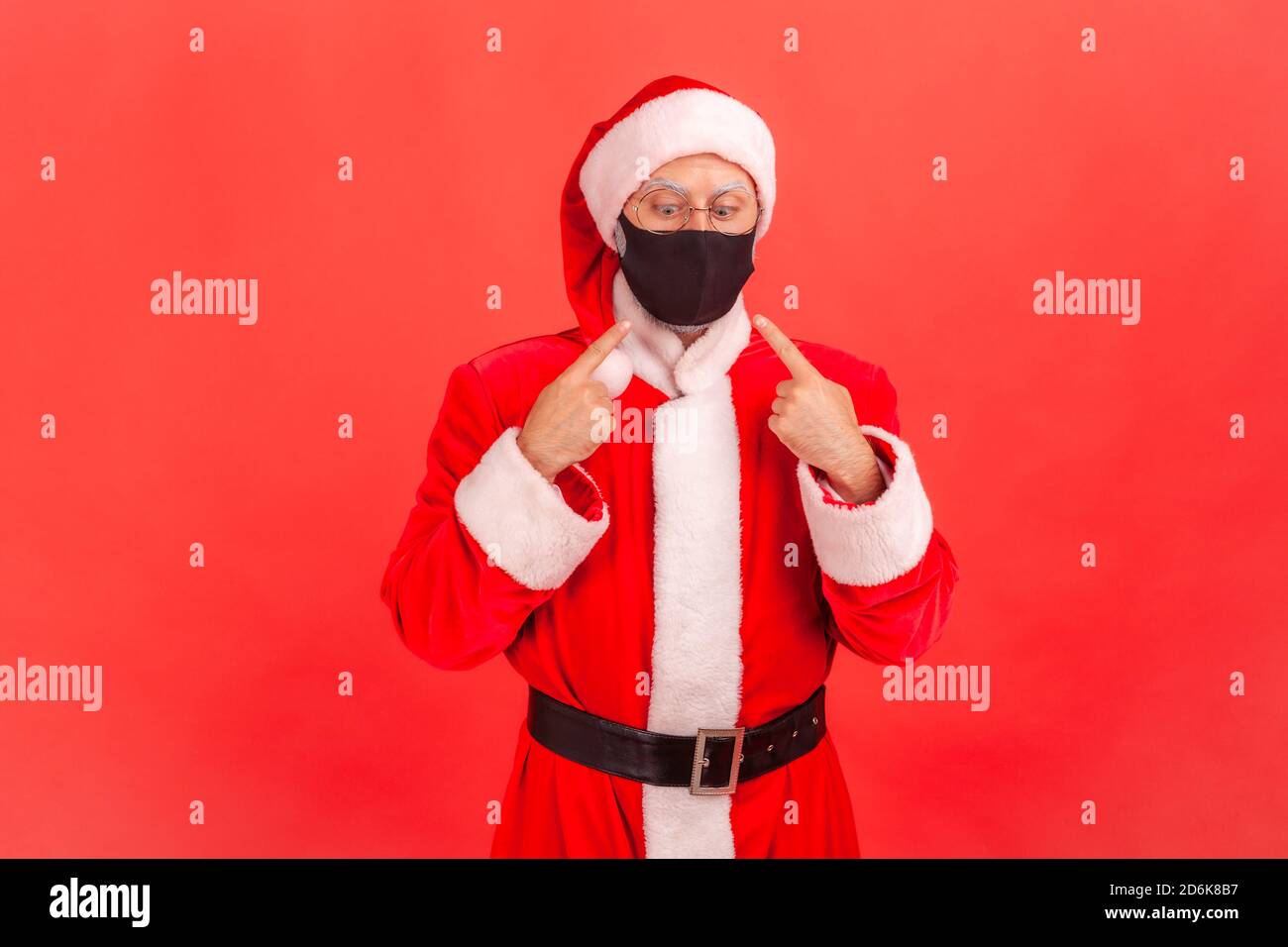 Scared funny man in santa claus costume pointing fingers and trying to look at protective hygienic mask on his face, tired of covid-19 epidemic. Indoo Stock Photo