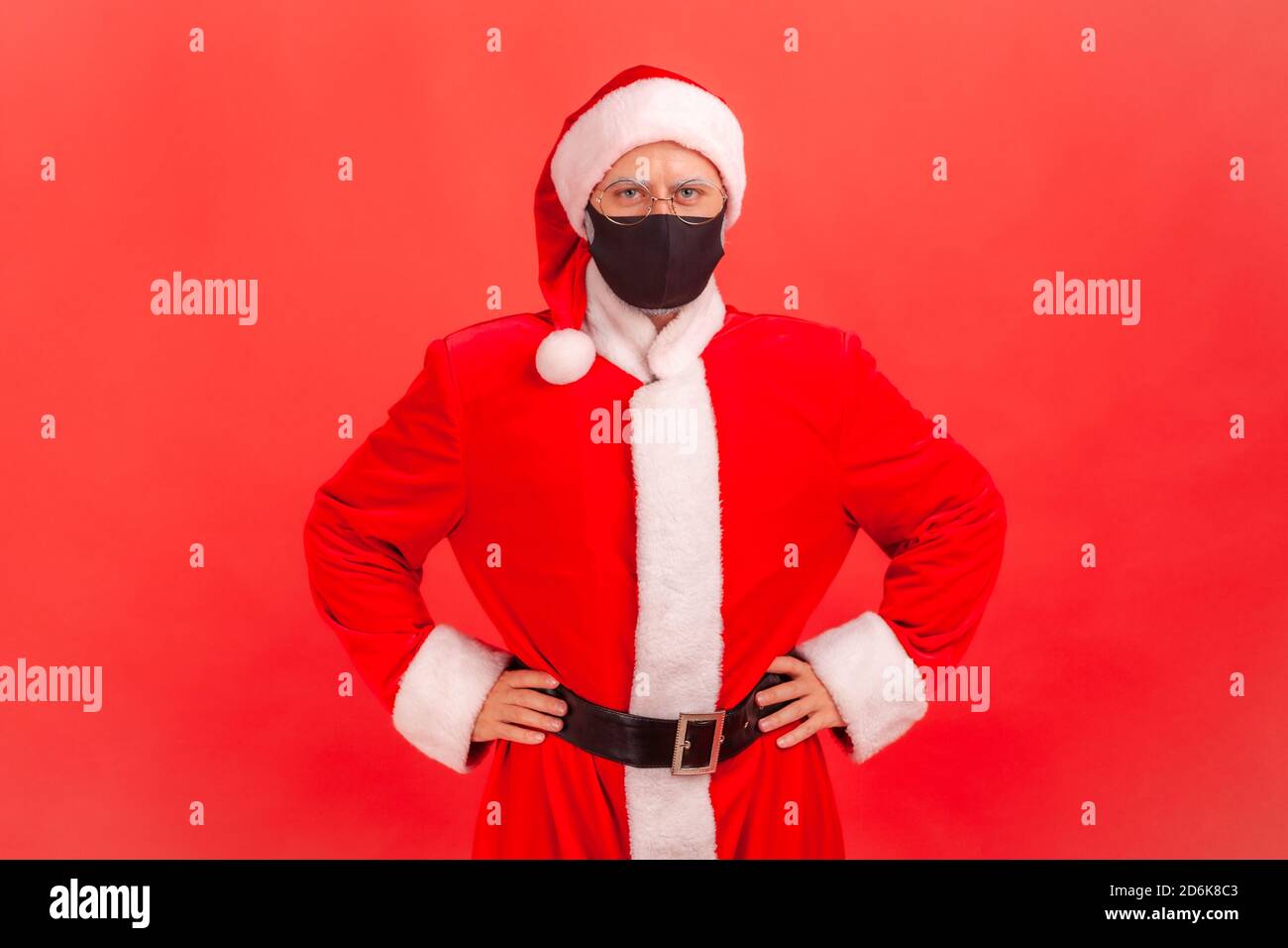 Serious concerned santa claus in eyeglasses wearing protective facial mask looking at camera holding hands akimbo, quarantine on holidays. Indoor stud Stock Photo