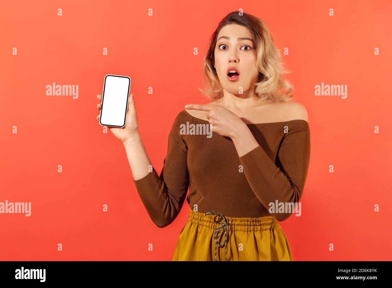Shocked surprised woman pointing finger at smartphone with white empty screen, looking with amazed expression, online app. Indoor studio shot isolated Stock Photo