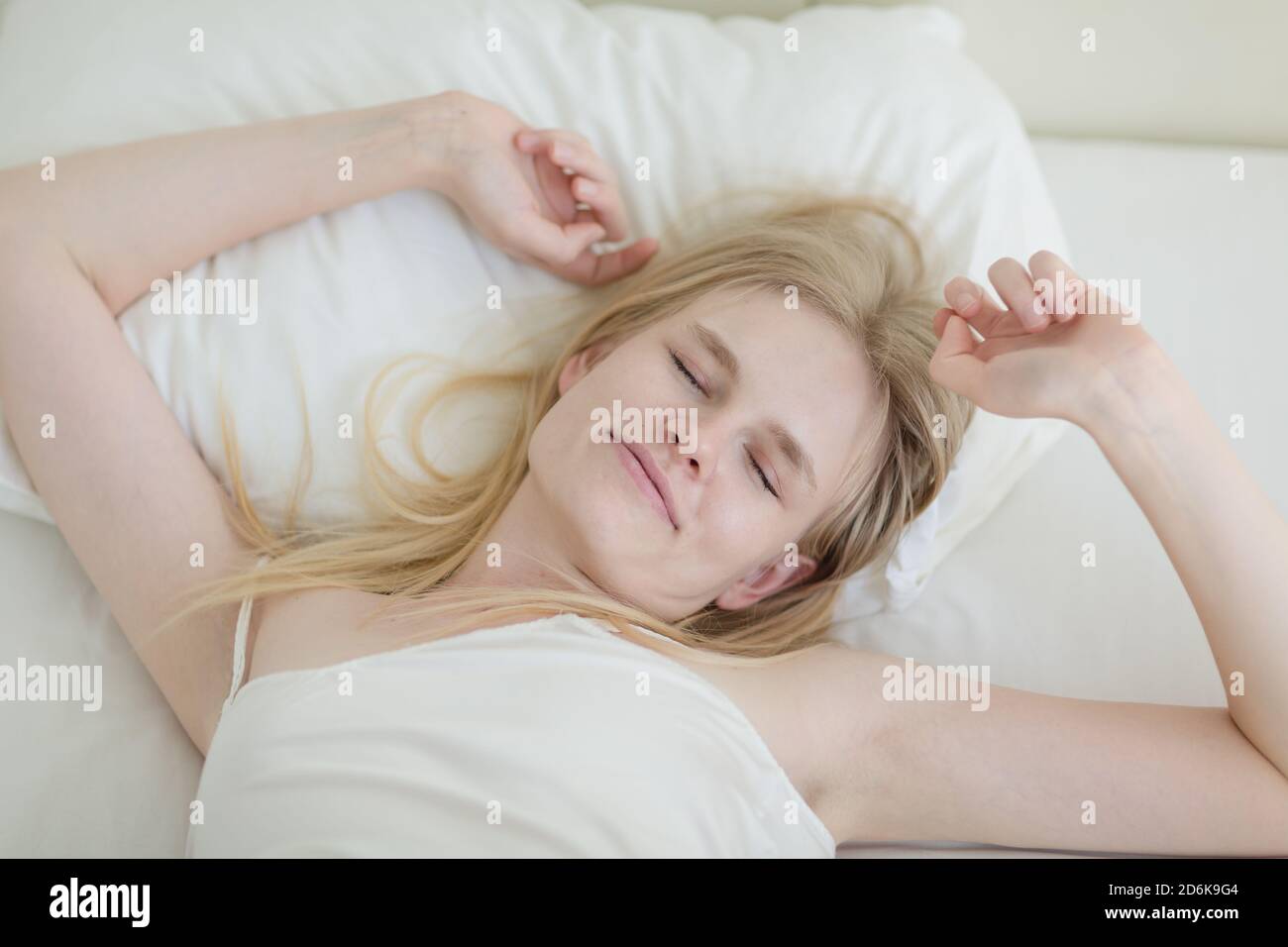 Beautiful young woman in bed waking up while stretching her arms in the morning relaxed from a good night's sleep. Stock Photo
