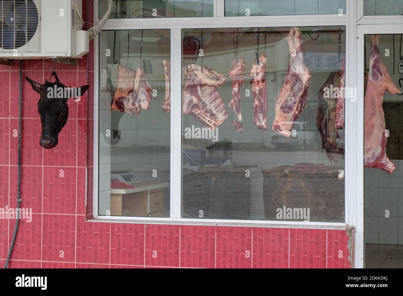 View through dirty blurred window glass to fresh meat carcasses hanging and severed head on hook in street butchers shop in caucasus village market. Stock Photo