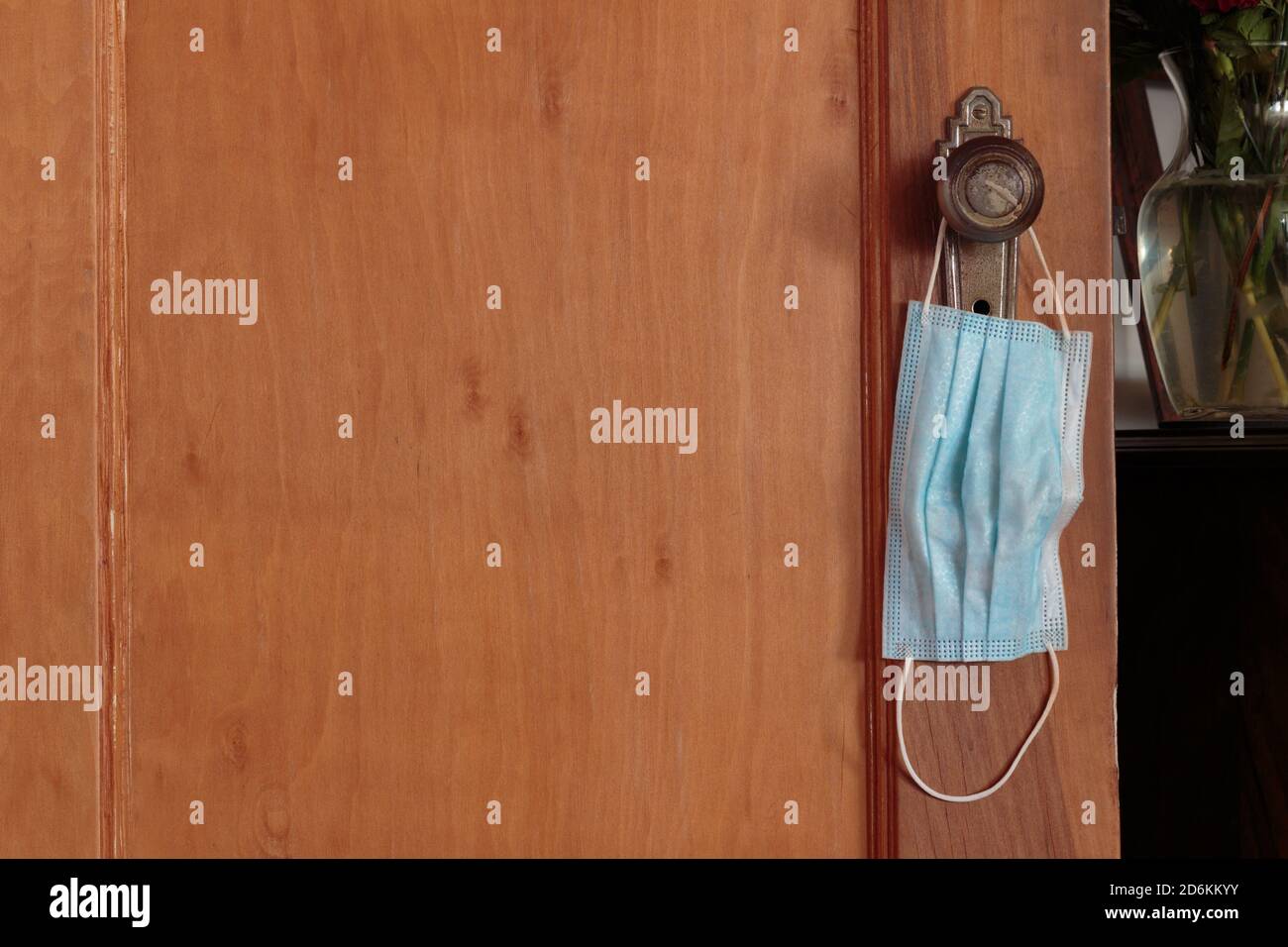 a blue face mask hanging from a door knob on a wooden door next to a table, illustrating the intrusion of face masks in our lives during the pandemic Stock Photo