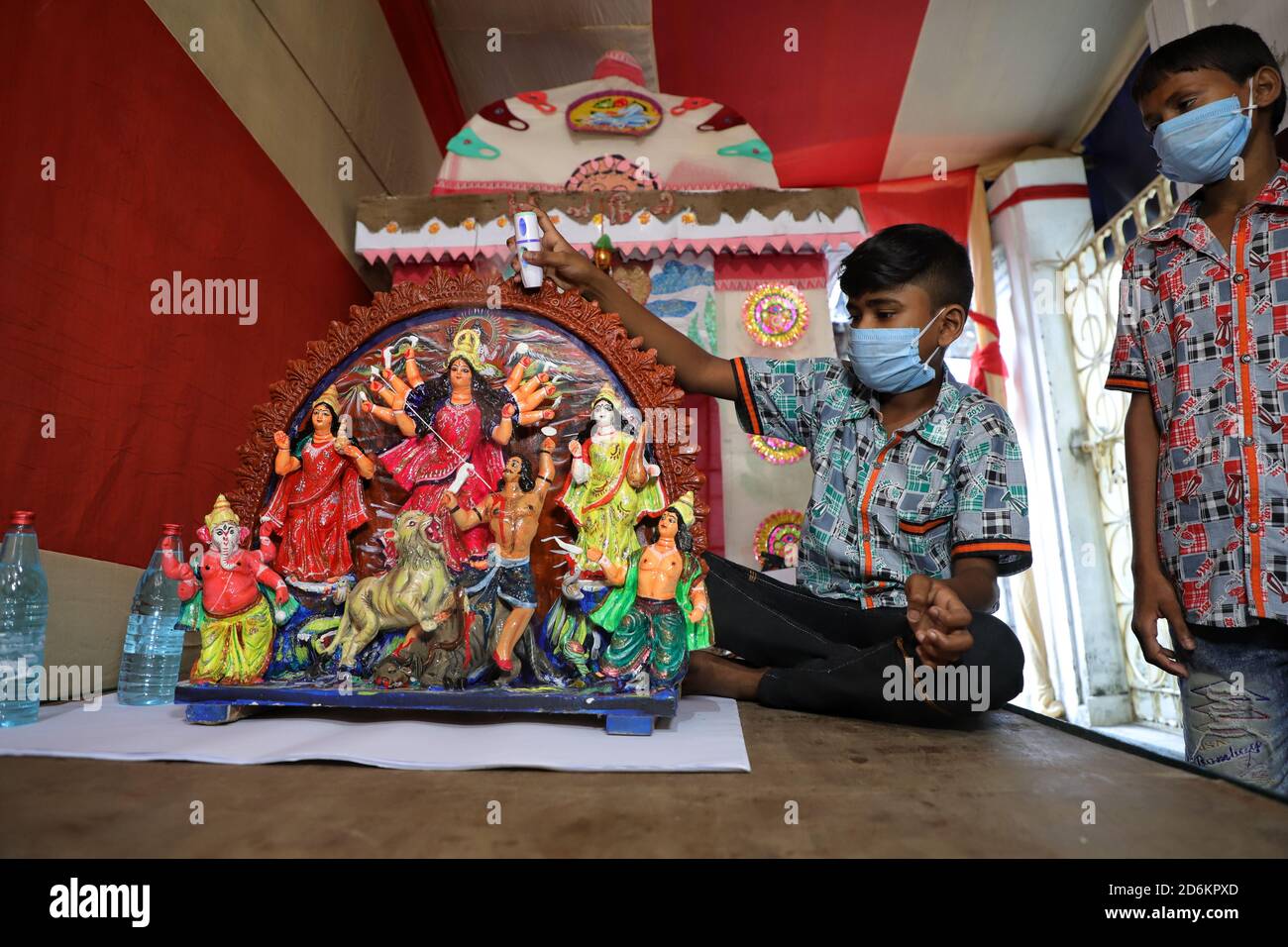 A boy wearing a facemask is seen seated and spraying a disinfectant to his 1.5 feet tall clay idol of goddess Durga in a pandal prior to Durga puja Festival amidst Covid-19 pandemic. Stock Photo