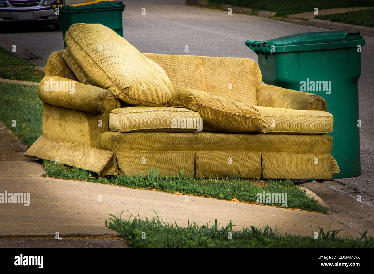 Old yellow couch with cushions sits on a curb in residential area next to a recycling cart waiting for bulk waste hauler pickup on trash day Stock Photo