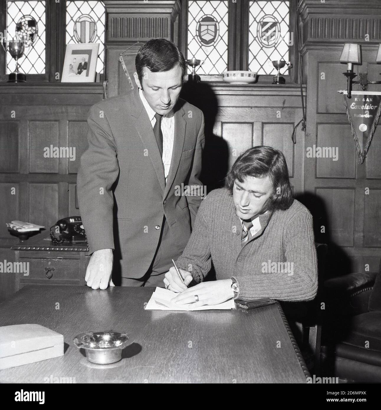 1970s, historical, a footballer in a club boardroom signing forms with his manager standing beside him, South East London, England, UK. Hanging on a light fitting is a pendant from SBV Excelsior, a football team in Rotterdam, Netherlands. Stock Photo