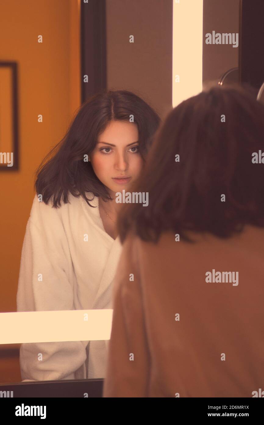 Girl with short dark hair looks at herself in a mirror, wearing a white bathrobe; she looks serious and/or thoughtful; self-reflection concept Stock Photo