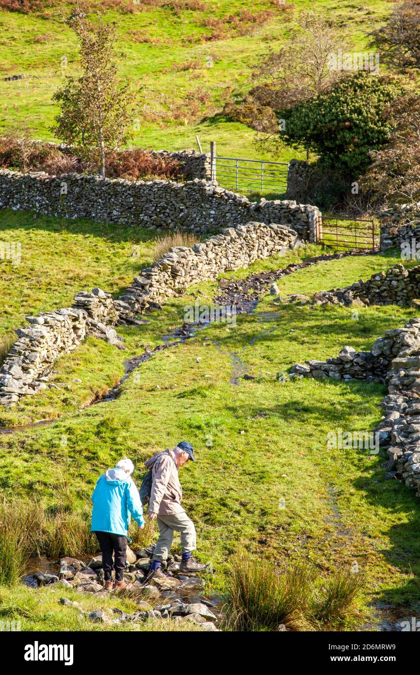 Elderly man and woman  pensioners senior citizens rambling in the north Yorkshire Dales crossing a stream / water in the hills above Kirkby Lonsdale Stock Photo