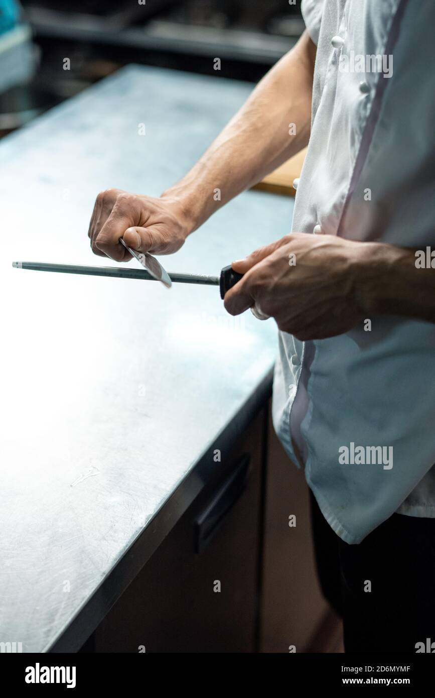 Hands of chef of restaurant in white uniform sharpening knife over kitchen table Stock Photo