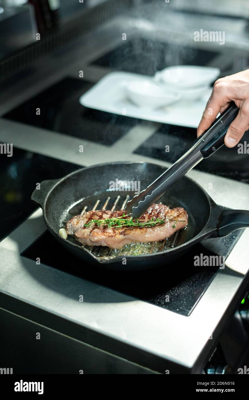 Hand of chef frying piece of beef and adding fresh aromatic herbs to the meat Stock Photo