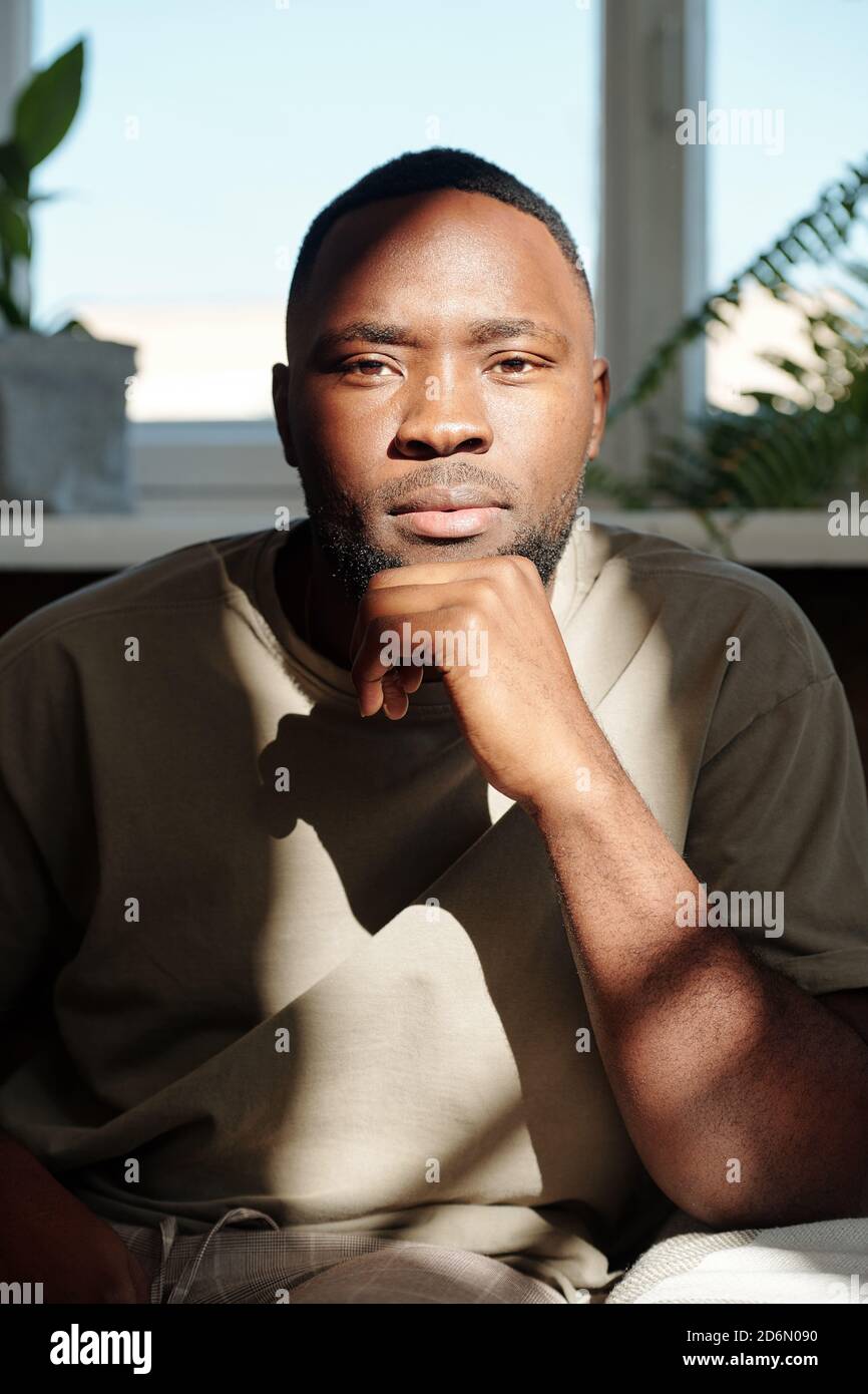 Young serious man of African ethnicity looking at you while sitting by window Stock Photo