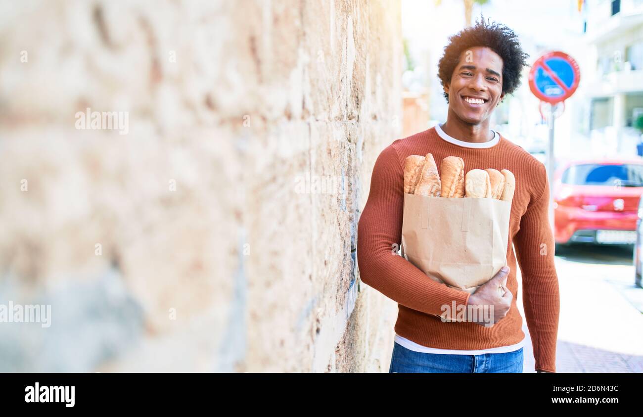 Young handsome african american man smiling happy. Standing with smile on face holding paper bag with bread walking at town street. Stock Photo