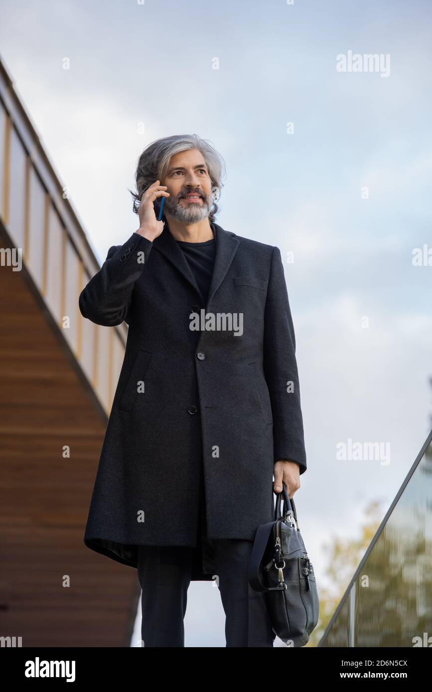 Confident grey-haired businessman in black coat making appointment by phone Stock Photo