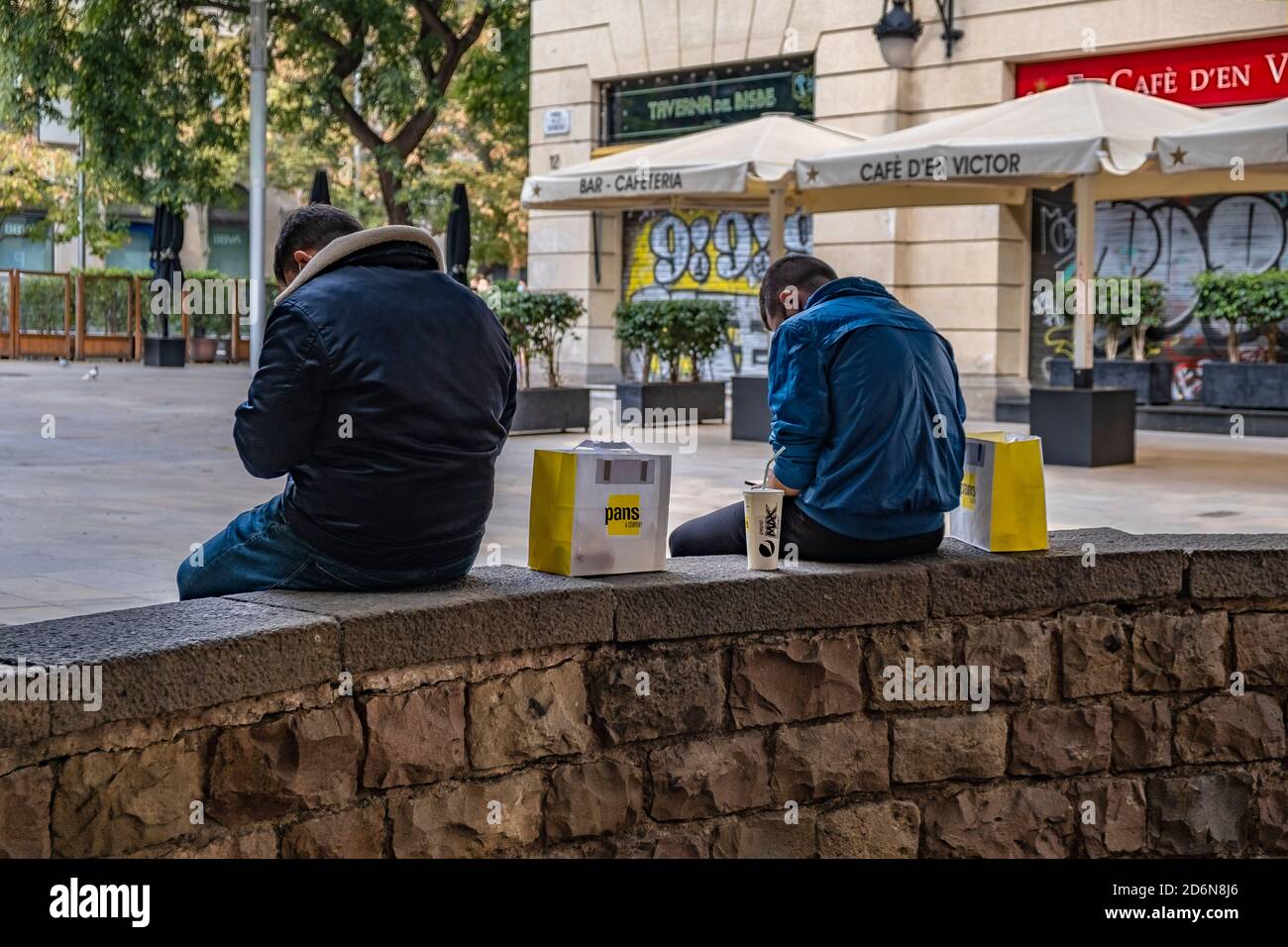 Two young men sitting on the street in front of a closed restaurant eat food prepared by the Spanish chain Pans and Company.The activity around take-out food has increased in Barcelona due to the decree of the Catalan Government to close the commercial activity of bars and restaurants for 15 days due to the increase in the contagion of Covid19. Stock Photo