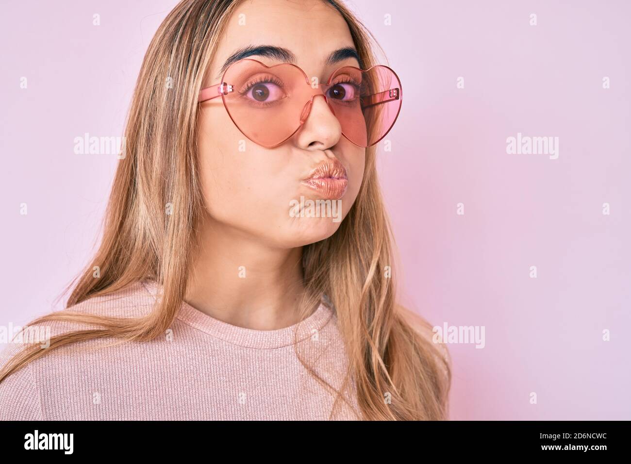 Young beautiful blonde woman wearing heart shaped sunglasses puffing cheeks with funny face. mouth inflated with air, catching air. Stock Photo