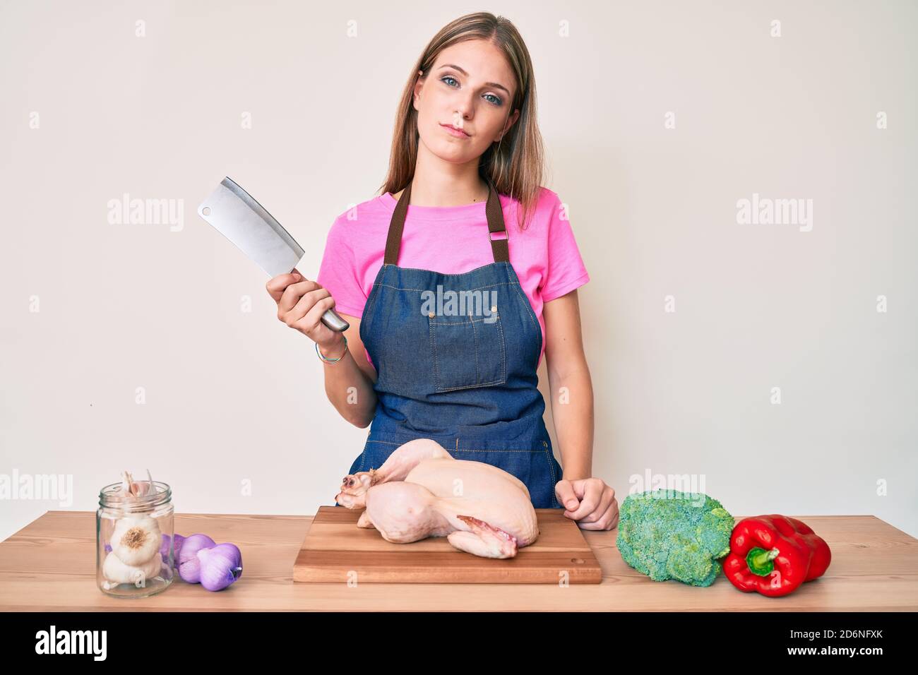 Young blonde girl wearing cook apron cooking chicken thinking attitude ...