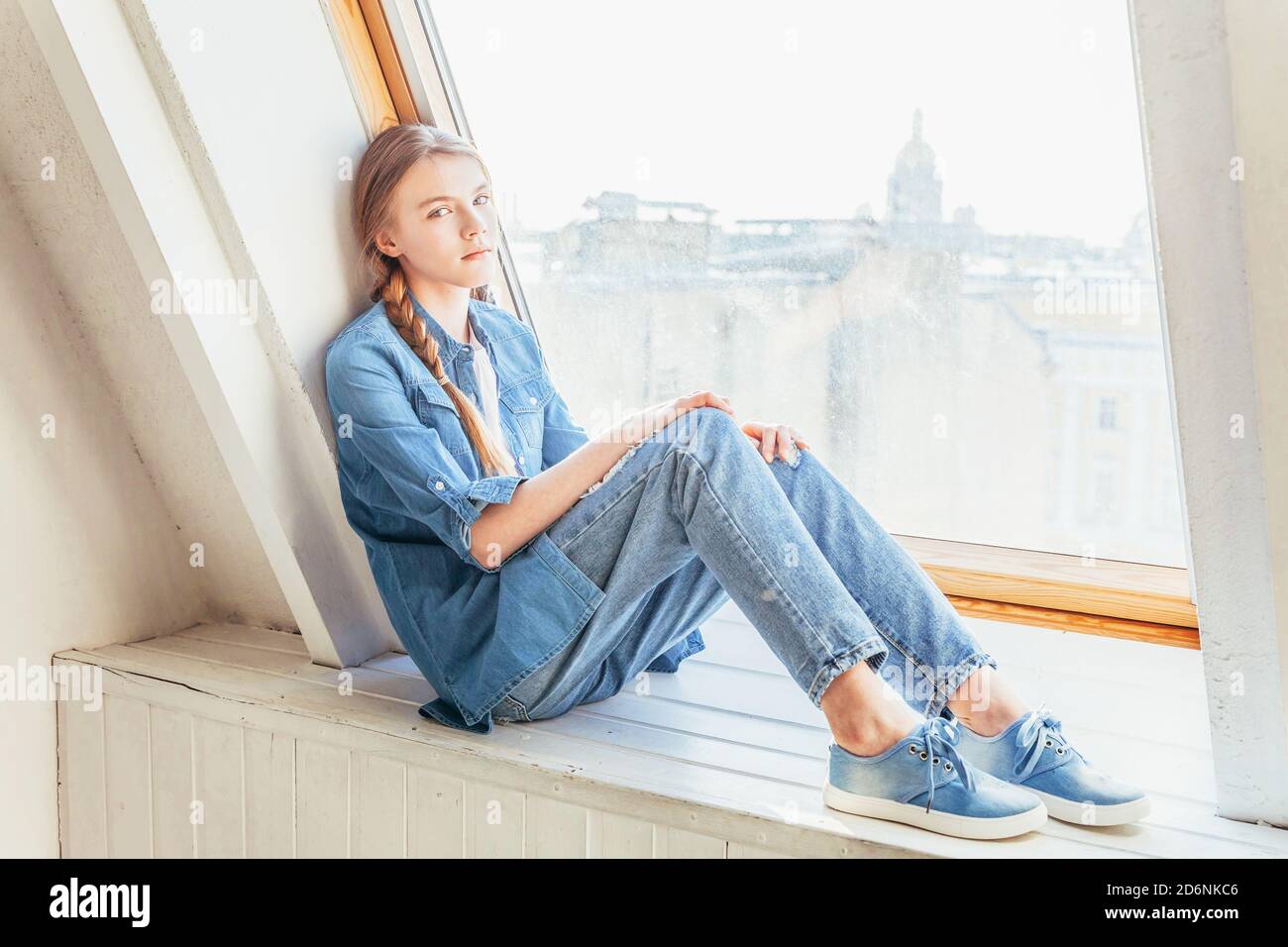 Young cute teenage girl in jeans, denim jacket and white T-shirt sitting on  window sill in bright light living room at home indoors and thinking.  Social distance jomo concept Stock Photo -