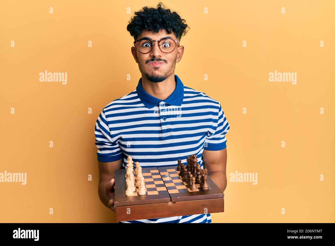 Young arab handsome man holding chess board wearing glasses puffing cheeks with funny face. mouth inflated with air, catching air. Stock Photo