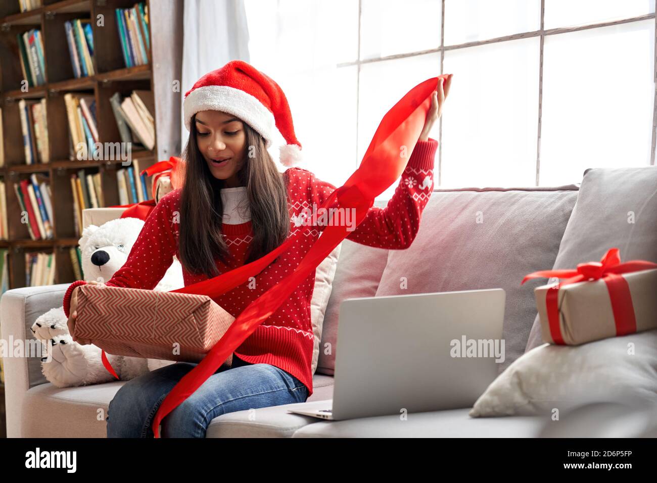 Excited indian child girl opening Christmas gift video calling family on laptop. Stock Photo