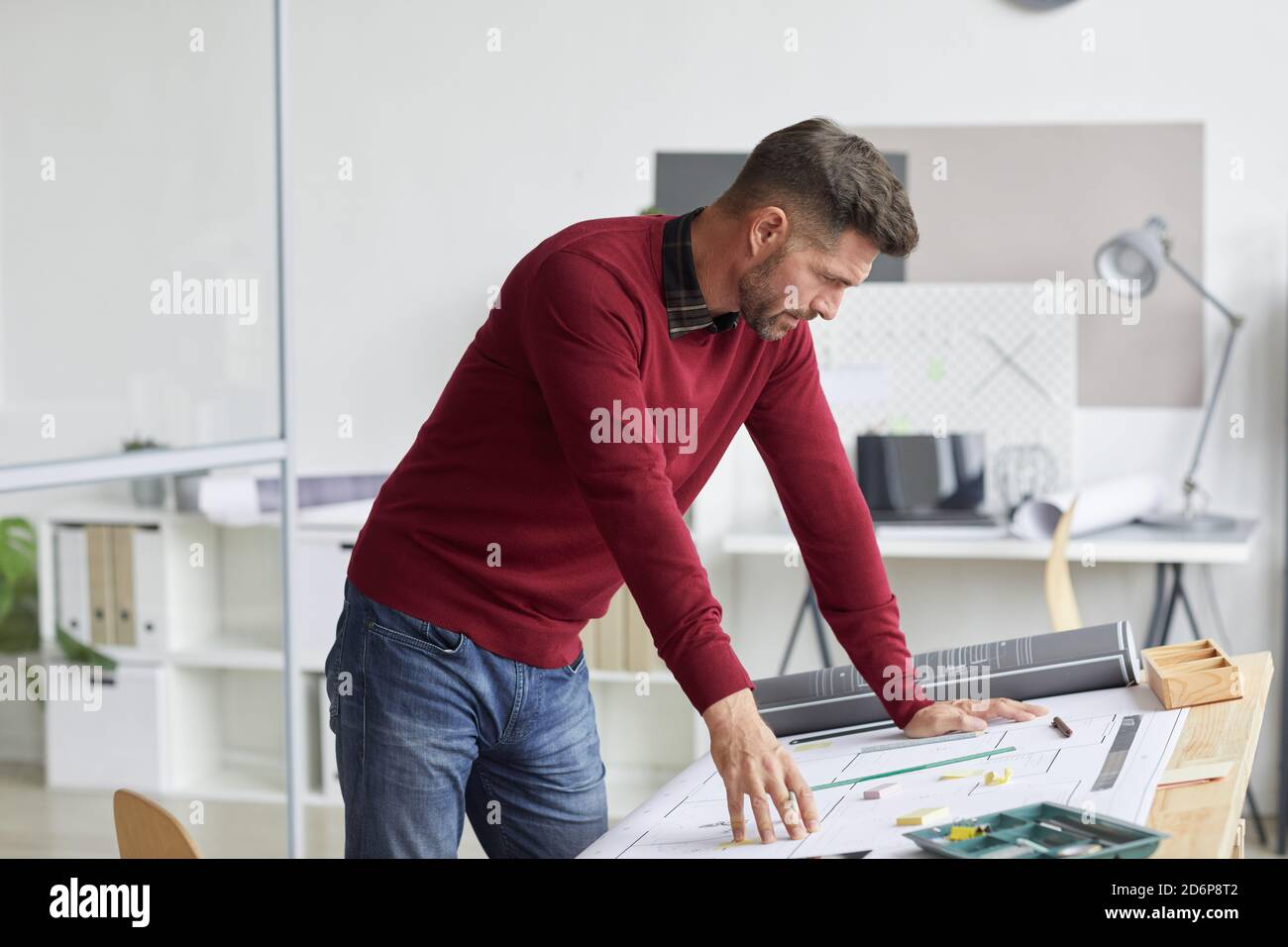 Side view portrait of bearded architect looking at blueprints while leaning on drawing table at workplace, copy space Stock Photo