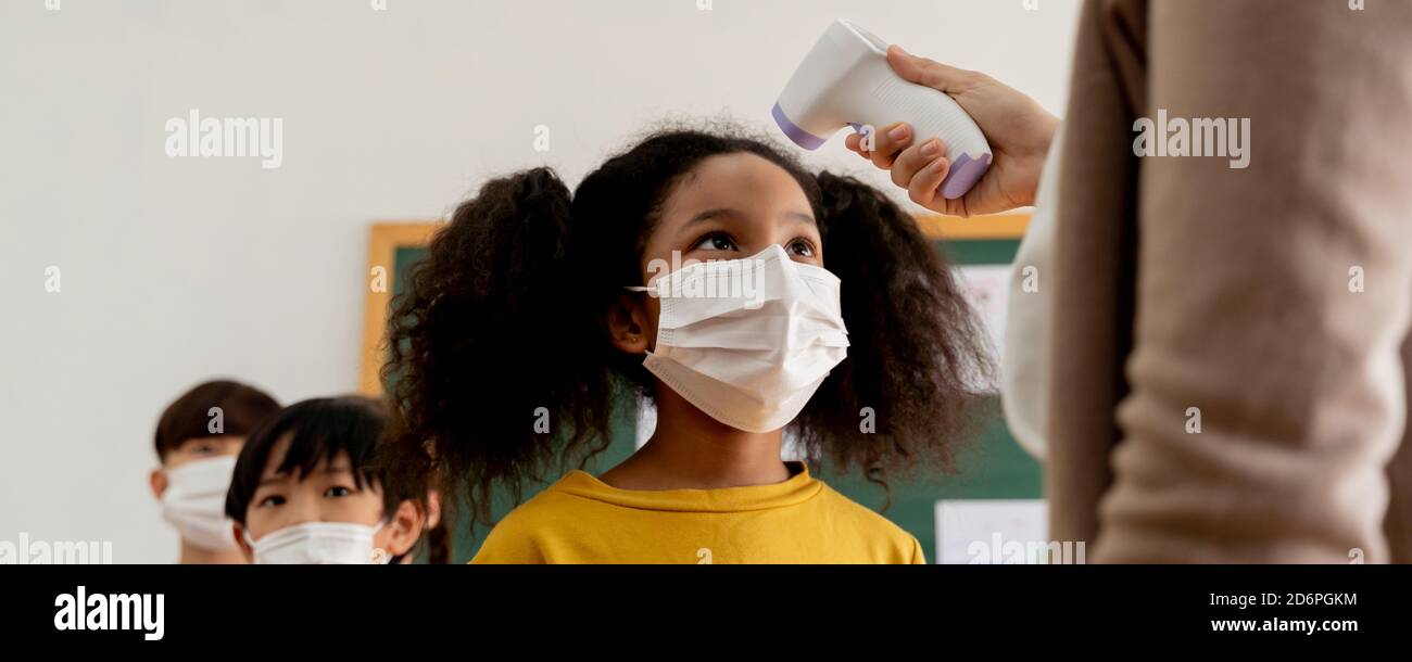 Banner group of diverse students in school building checked and scanned for temperature check. Elementary pupils are wearing face mask, line up before entering into classroom. Covid-19 school reopen Stock Photo