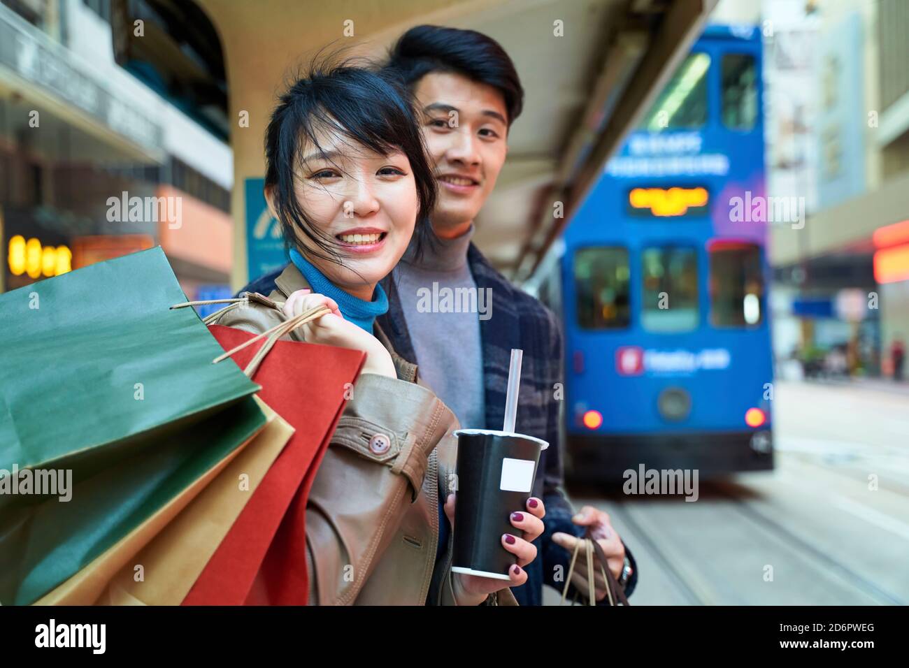 happy young asian couple waiting for bus in modern city with shopping bags in hand Stock Photo