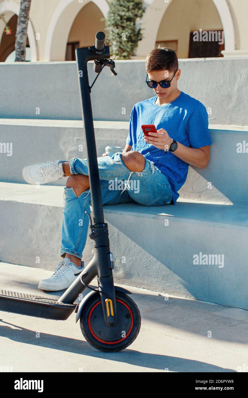 Young Caucasian man with smartphone sitting outdoor near electric scooter Stock Photo