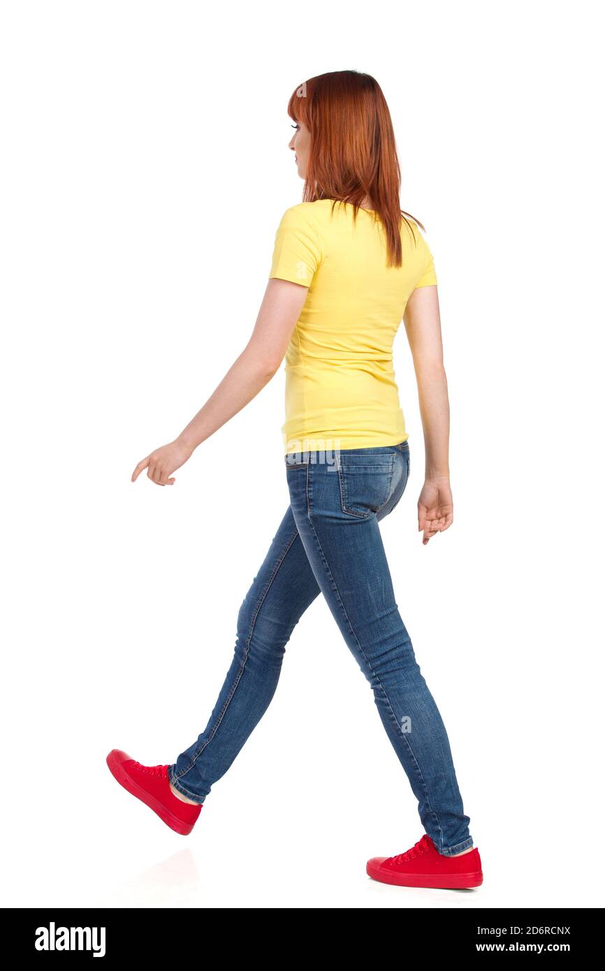 Walking young woman in yellow shirt, jeans and red sneakers. Side view. Full length studio shot isolated on white. Stock Photo
