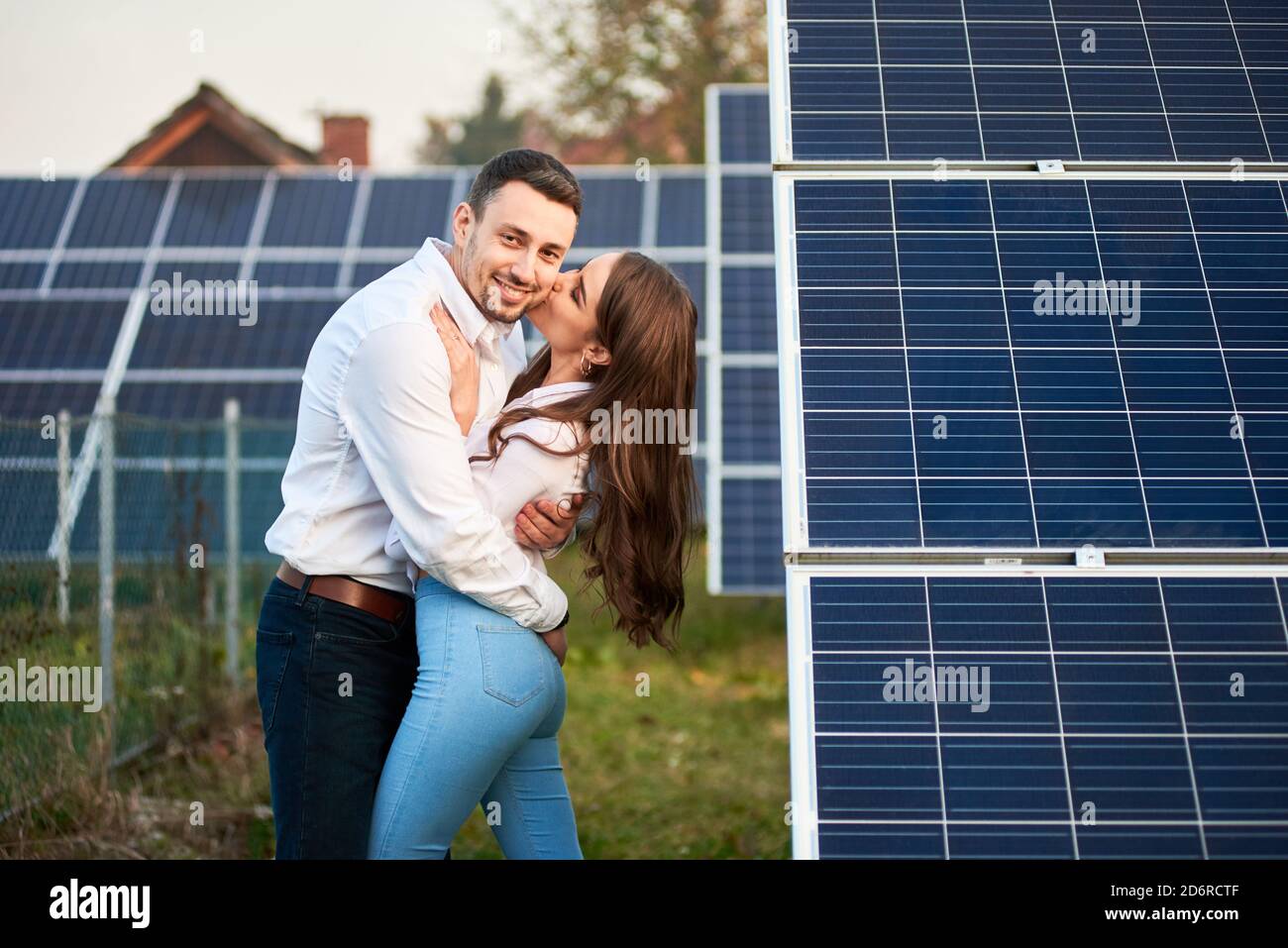 Happy couple are hugging against the background of a row of solar panels at a site near the house. Woman with long hair kisses a guy, man smiling to the camera. Solar energy concept image Stock Photo